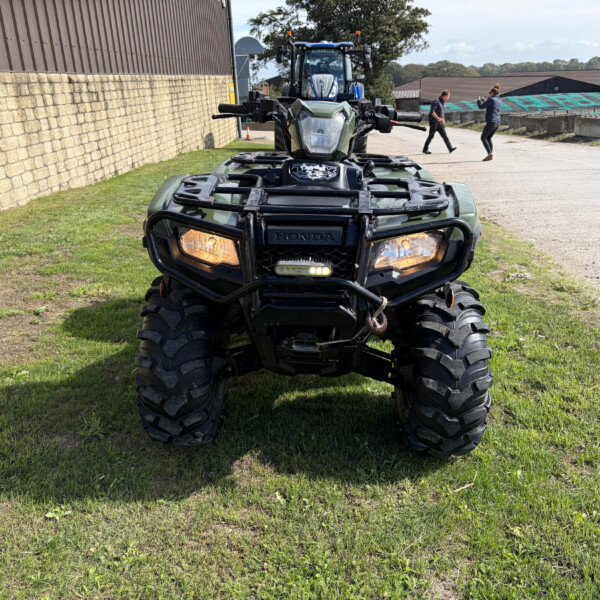 Front view of a green Honda ATV on grass, with a stone wall to the left and people near a blue tractor in the background.