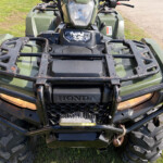 Front view of a green Honda ATV with a metal cargo rack, headlights, and muddy tires on grass.