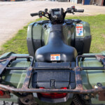 Back view of a green ATV with a rusted metal cargo rack, grass and industrial area in the background, warning stickers on the side panel visible.