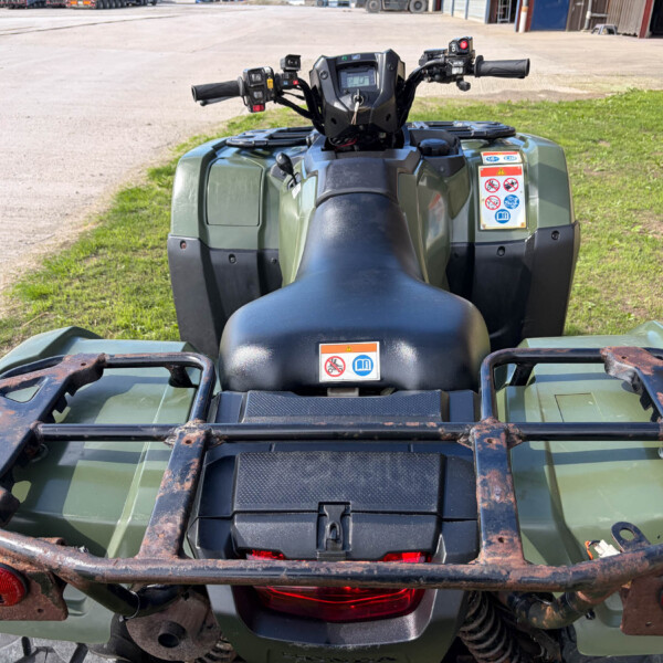 Back view of a green ATV with a rusted metal cargo rack, grass and industrial area in the background, warning stickers on the side panel visible.