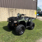 Green Honda ATV parked on a grassy area beside a beige brick wall and brown metal building.