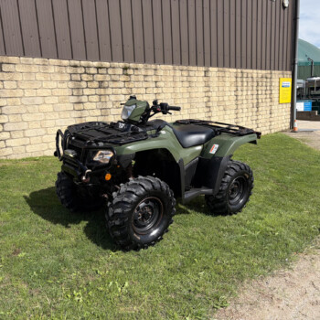 Green ATV with cargo racks parked on grass beside a stone wall and brown metal-clad building.