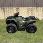 Green ATV with large tread tires parked on grass, in front of a stone retaining wall and brown metal siding.