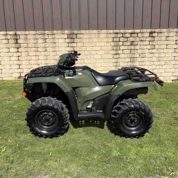 Green ATV with large tread tires parked on grass, in front of a stone retaining wall and brown metal siding.