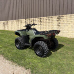 Green ATV parked on grass beside a beige stone wall and brown metal building in the background.