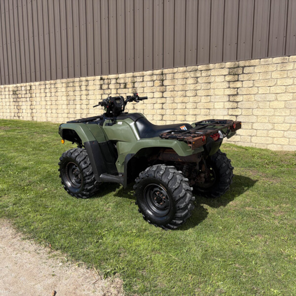 Green ATV parked on grass beside a beige stone wall and brown metal building in the background.