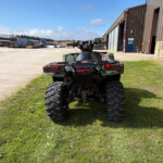 Rear view of a green ATV with large tires parked in a wide industrial yard, with warehouses and trailers in the background under a blue sky.