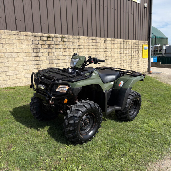 Green Honda ATV parked on a grassy area beside a beige brick wall and brown metal building.