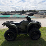 Close-up of a green ATV on grass with an industrial complex and cloudy sky in the background.