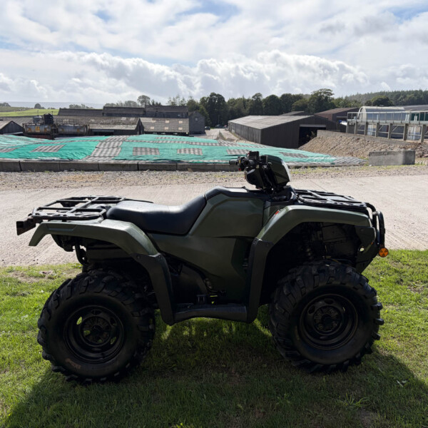 Close-up of a green ATV on grass with an industrial complex and cloudy sky in the background.