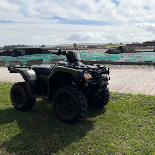 All-terrain vehicle (ATV) parked on grass in a rural industrial area, with low warehouses and green fields in the distance under a blue sky with clouds.