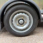 Close-up of a trailer wheel: black tire mounted on a silver steel rim with lug nuts, metal fender and trailer frame visible on the sides.