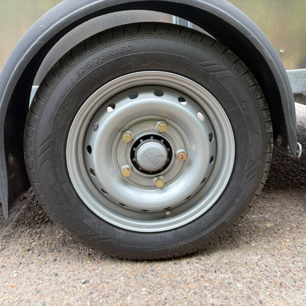 Close-up of a trailer wheel: black tire mounted on a silver steel rim with lug nuts, metal fender and trailer frame visible on the sides.