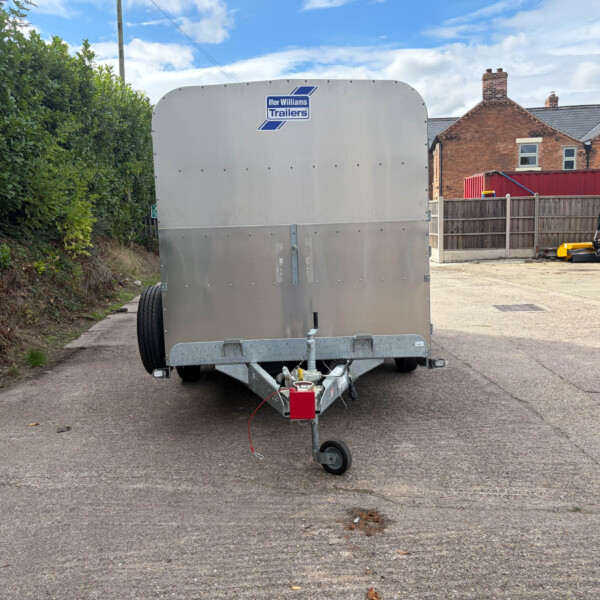 Silver Ifor Williams trailer parked on a paved lot beside a hedge, with red-brick houses and a yellow construction machine in the background.