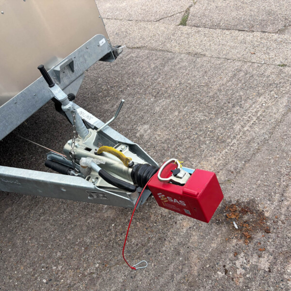 Trailer hitch assembly with a red battery box mounted to a galvanized frame on a rough asphalt surface, showing winch components and safety chains.