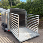 Open aluminum trailer with side rails and lowered ramp in a parking lot beside a hedge and a storefront sign behind it.