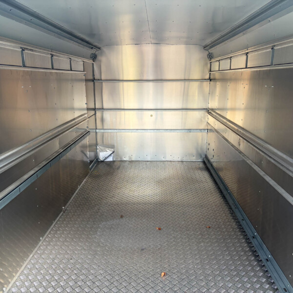 Interior of a metallic empty cargo trailer with diamond-plate floor and riveted panels along the walls, viewed from the end.