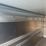 Interior of a stainless steel trailer or cabinet with riveted panels, rail tracks, and a diamond-plate floor; empty shelving along the walls.