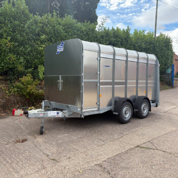Silver enclosed utility trailer parked on a street beside a tall hedge, with a hitch and spare wheel visible.