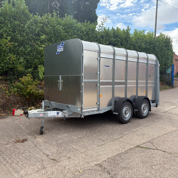 Silver enclosed utility trailer parked on a street beside a tall hedge, with a hitch and spare wheel visible.