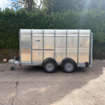 Silver metal cargo trailer parked on an asphalt surface with a dense green hedge and blue sky in the background.