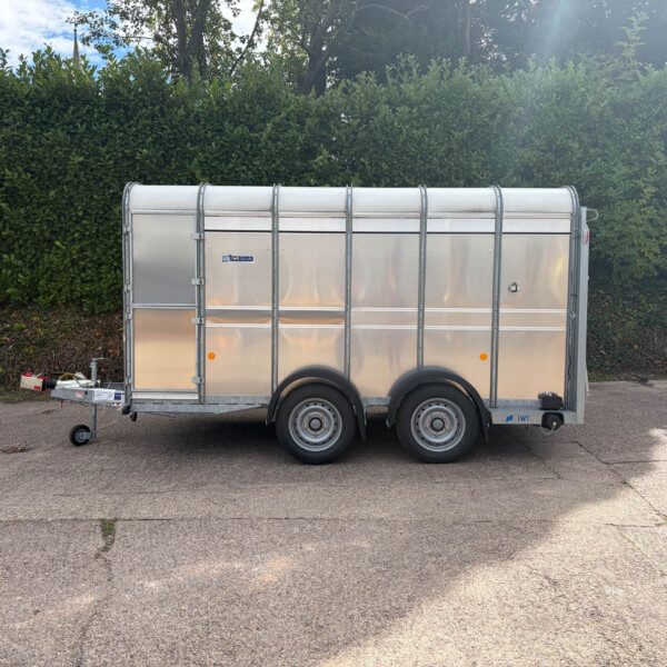 Silver metal cargo trailer parked on an asphalt surface with a dense green hedge and blue sky in the background.