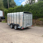 Silver metal livestock trailer with rounded roof, parked in a lot beside a tall green hedge and a white storefront building.