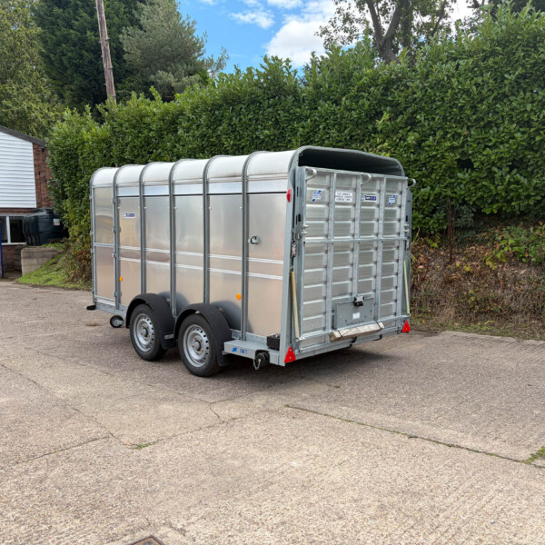 Silver metal livestock trailer with rounded roof, parked in a lot beside a tall green hedge and a white storefront building.