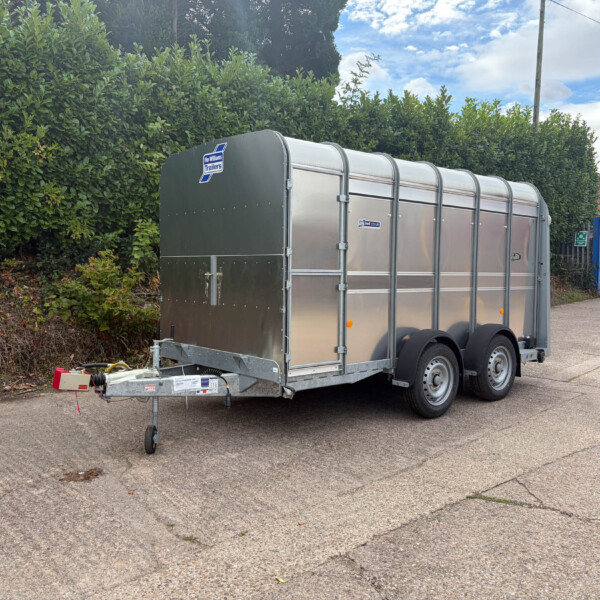 Silver enclosed trailer with curved roof parked on a street beside a tall green hedge; visible hitch and equipment on the ground in front of it
