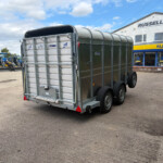 Metal livestock trailer attached to a dual-axle cart in a dealership lot, with tractors lined up on the left and a Russell’s store sign in the background.
