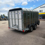 Rear view of a large metal livestock trailer on a paved lot, with blue tractors on the left and a Russell's storefront in the background.