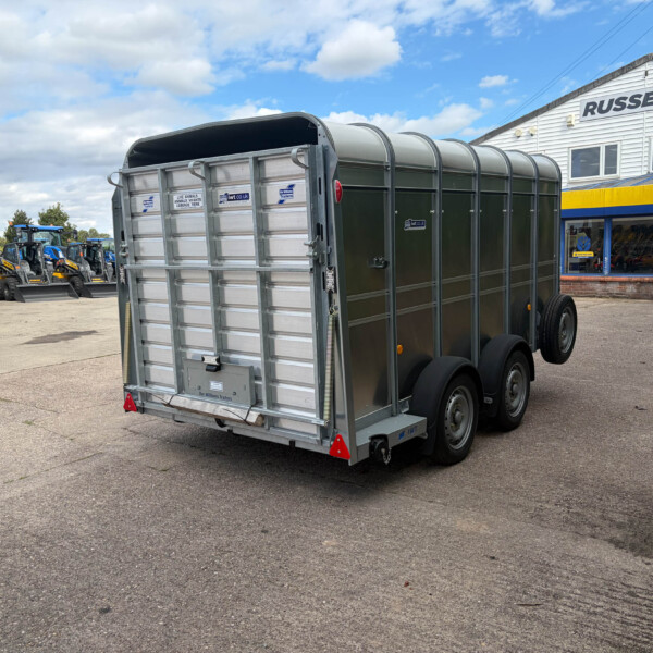 Rear view of a large metal livestock trailer on a paved lot, with blue tractors on the left and a Russell's storefront in the background.
