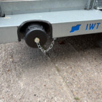 Close-up of a metal trailer bumper with a black side-mounted wheel, safety chains, and a blue TWT logo on a gravel surface.