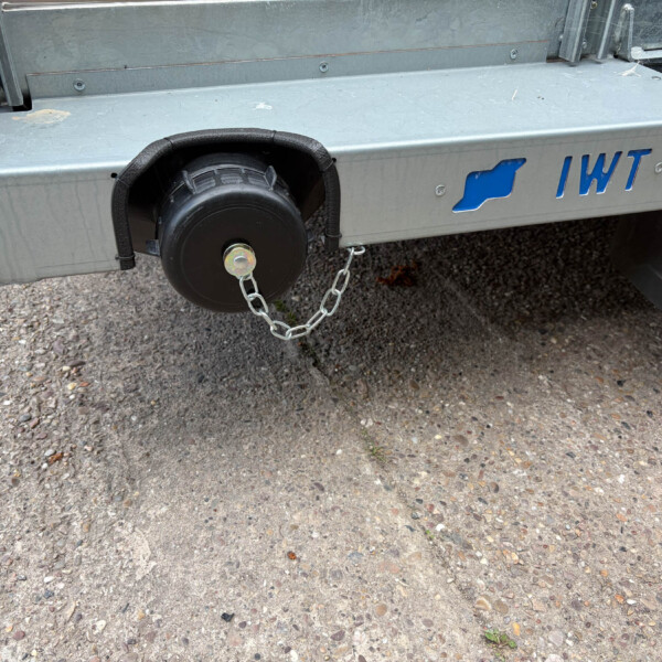 Close-up of a metal trailer bumper with a black side-mounted wheel, safety chains, and a blue TWT logo on a gravel surface.
