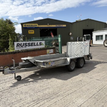 Farm equipment yard with a green corrugated warehouse, a Russell’s sign, and a blue New Holland Agriculture sign; a blue tractor and trailer are visible on the right side of the image.