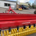Red agricultural implement with yellow roller tines, branded Pöttinger, parked in an outdoor equipment yard beside industrial buildings.