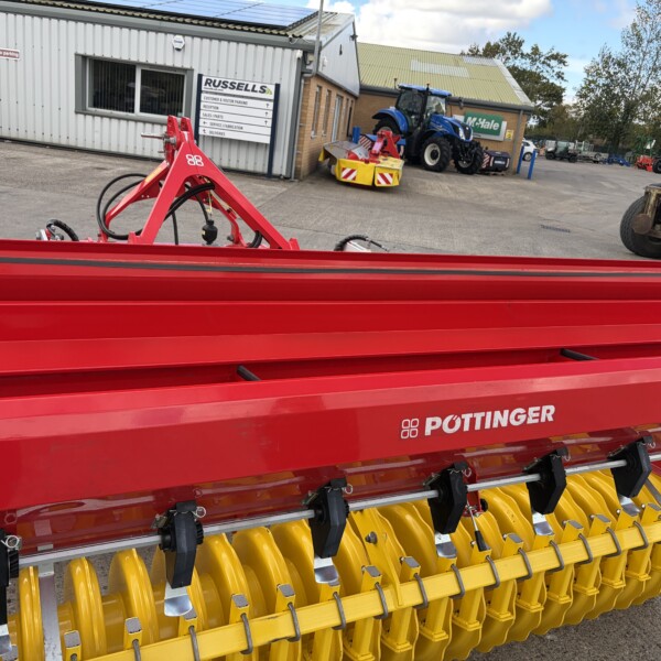 Red agricultural implement with yellow roller tines, branded Pöttinger, parked in an outdoor equipment yard beside industrial buildings.