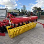 Red and yellow agricultural seed drill parked in a yard with tractors and a sign in the background, under a blue sky.