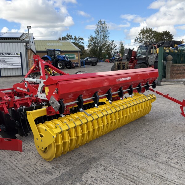 Red and yellow agricultural seed drill parked in a yard with tractors and a sign in the background, under a blue sky.