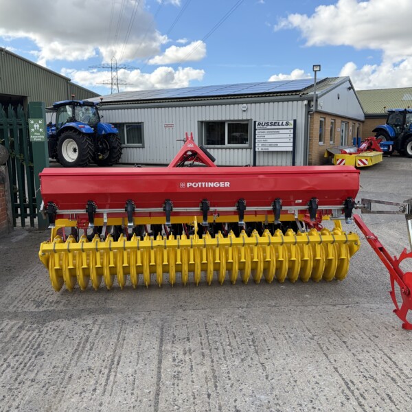 Yellow Pottinger seed drill in the foreground with red support, in a farm equipment yard among blue tractors and gray metal buildings in the background.