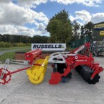 Farm machinery display outside a New Holland Agriculture service building, with a Russell banner in the background and red and yellow implements lined up.