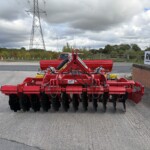 Red farming cultivator with multiple black disc harrows, parked in an outdoor lot beside a brick wall and a Russell sign.