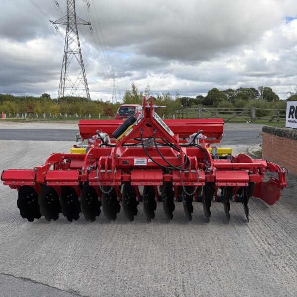 Red farming cultivator with multiple black disc harrows, parked in an outdoor lot beside a brick wall and a Russell sign.