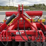 Close-up of a red agricultural implement with hydraulic hoses and metal linkage, outdoors on a farm driveway.