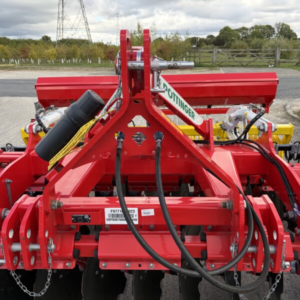 Close-up of a red agricultural implement with hydraulic hoses and metal linkage, outdoors on a farm driveway.