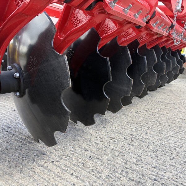 Red agricultural plow with curved black disks laid out in a row on a paved surface, showing metal bolts and chains nearby.
