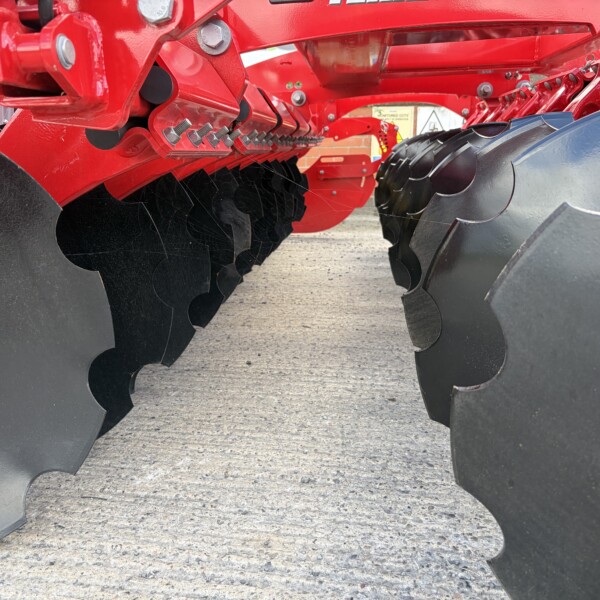 Close-up of a red disc harrow with scalloped black blades and metal bolts used for soil tilling.
