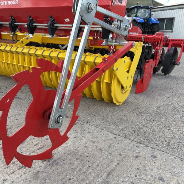 Red and yellow agricultural drill/air seeder with a red frame, yellow discs, and metal ladders, parked on a paved lot; a blue tractor is in the background.