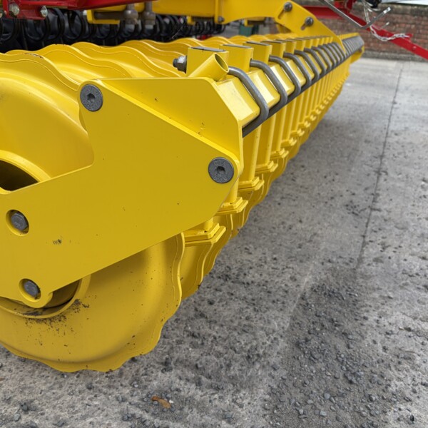 Close-up of yellow agricultural/industrial machinery with linked metal segments and bolts, red frame visible in background.