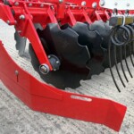 Close-up of a red farming implement with serrated black discs and coiled hoses on a gravel surface